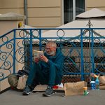 Homeless man reading a book on a city street in Kraków, sitting by a blue railing with a cardboard sign asking for help.
